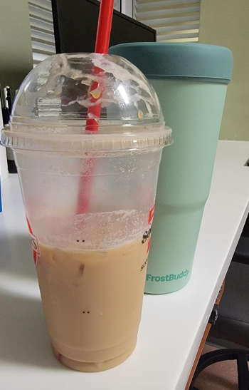 Two cups sit on a table; one is a disposable cup with a drink and straw, next to a reusable tumbler.