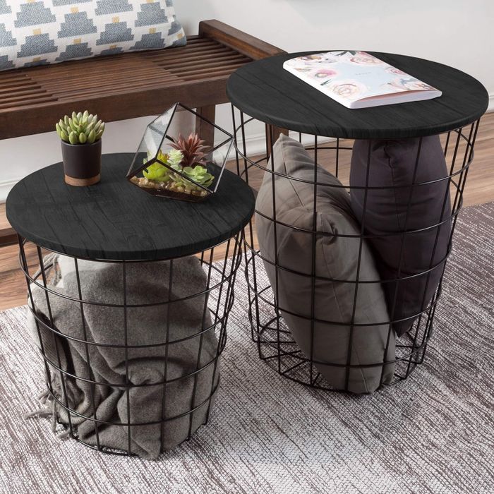 Two round, black wireframe side tables with wooden tops, one holding a plant and an open magazine, while the other features a blanket and pillow.