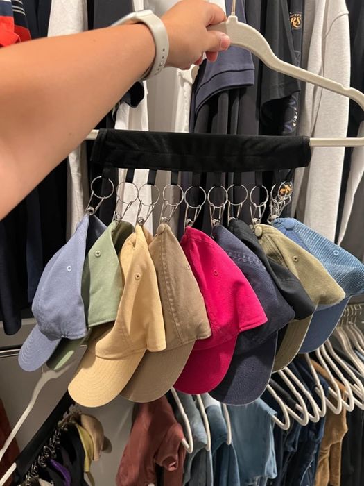 A hand grips a hanger showcasing nine baseball hats in a variety of styles and materials, neatly arranged side by side in a retail setting.