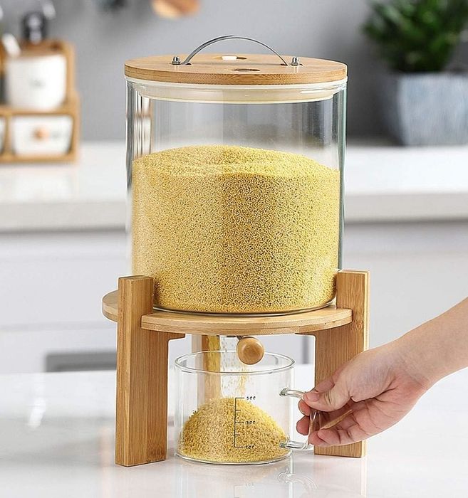 A hand pouring millet grains from a stylish glass and bamboo container into a measuring cup on a kitchen counter