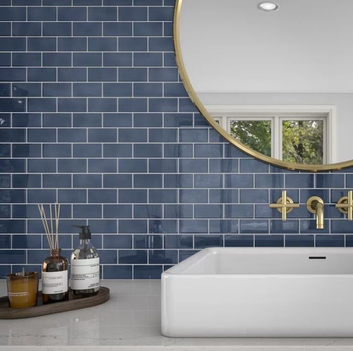 Modern bathroom featuring a round mirror, navy blue subway tile backsplash, a white rectangular sink, and gold fixtures. Essential items rest on a tray by the sink.