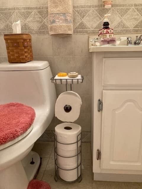 A bathroom featuring a toilet, tissue holder, and sink, with extra toilet paper rolls and decorative soaps neatly arranged on a metal rack.
