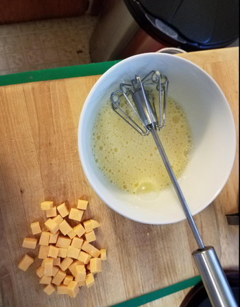 A whisk placed next to a bowl of eggs that have been beaten together.