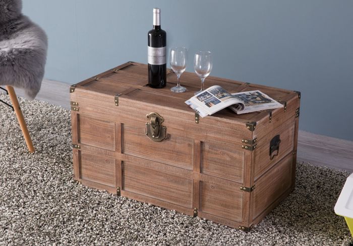 An antique-style wooden trunk used as a coffee table, adorned with a wine bottle, two glasses, and an open magazine, all resting on a plush rug