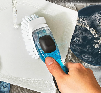 A hand holding a scrub brush, cleaning a white plate under running water