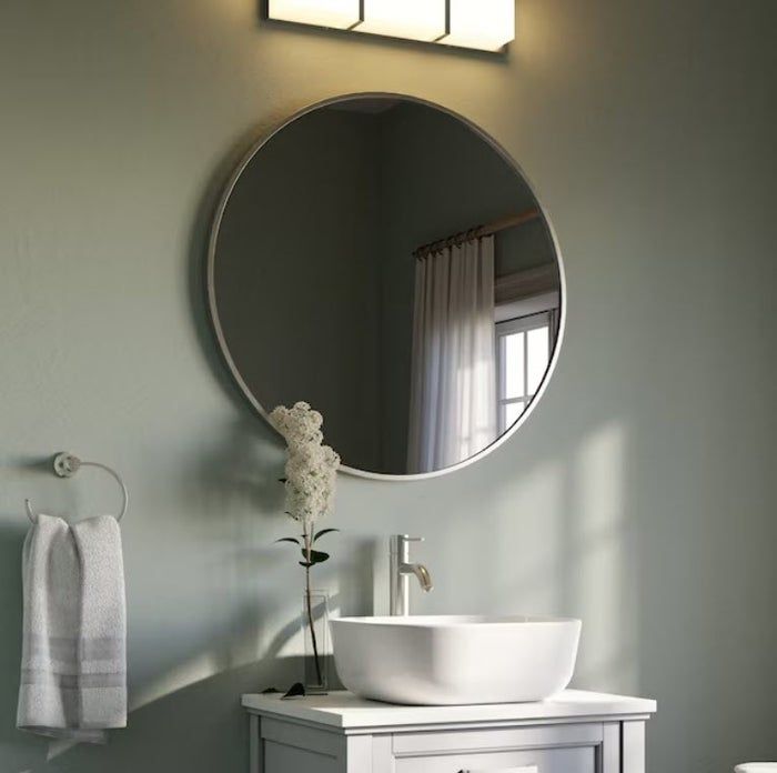 Modern bathroom featuring a round mirror, white sink, towel hung on a hook, and a vase with white flowers on the counter. A light fixture is placed above the mirror.