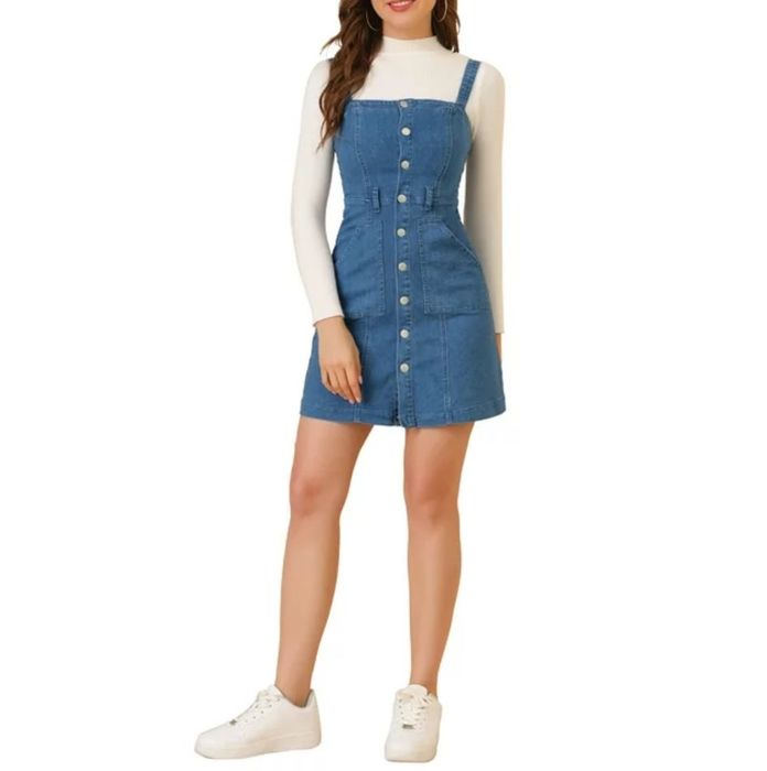 A model wearing a denim overall dress paired with a white long-sleeve top and white sneakers