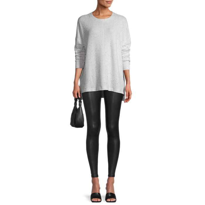 A person dressed in a long-sleeve top, black leggings, and black heels, holding a small bag. Their face is not shown.