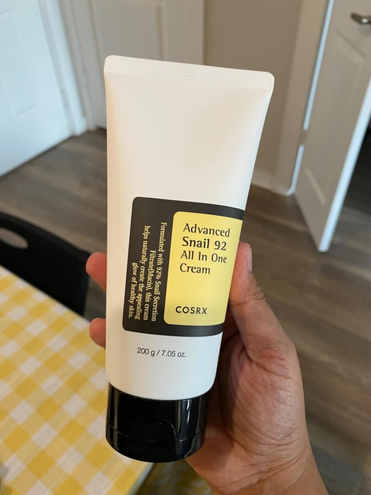Person holding a tube of COSRX Advanced Snail 92 All In One Cream, 200g, on a checkered tablecloth.