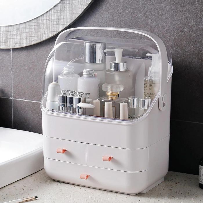 A clear-lidded organizer with a handle, packed with various skincare and beauty products, such as jars and bottles. Beneath it, two drawers with pink handles provide extra storage.