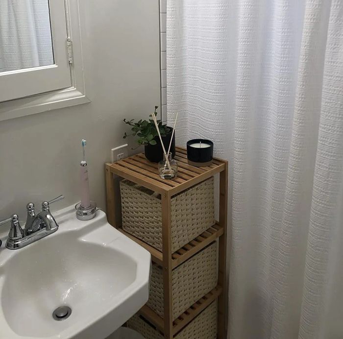 A bathroom featuring a white shower curtain, sink, and a wooden shelf holding wicker baskets, a small plant, reed diffuser, and a candle.