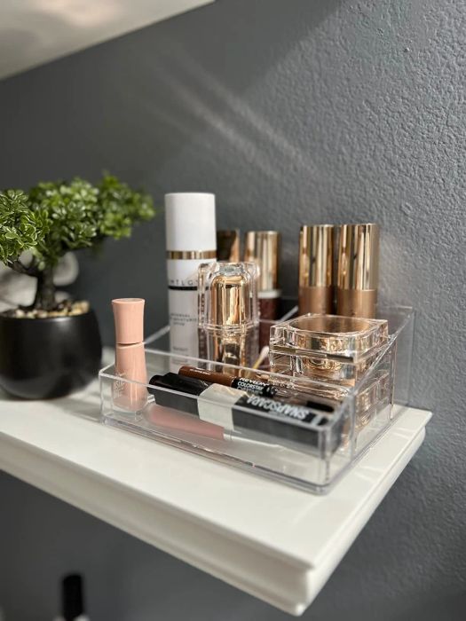 A transparent plastic organizer filled with various makeup items like lipsticks, foundation, setting spray, and eyeliners, placed on a shelf beside a small potted plant