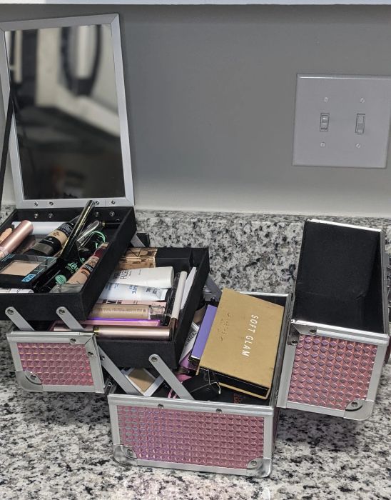 An open makeup case resting on a granite counter, filled with a variety of makeup items such as mascara, eyeshadow palettes, and lip products.
