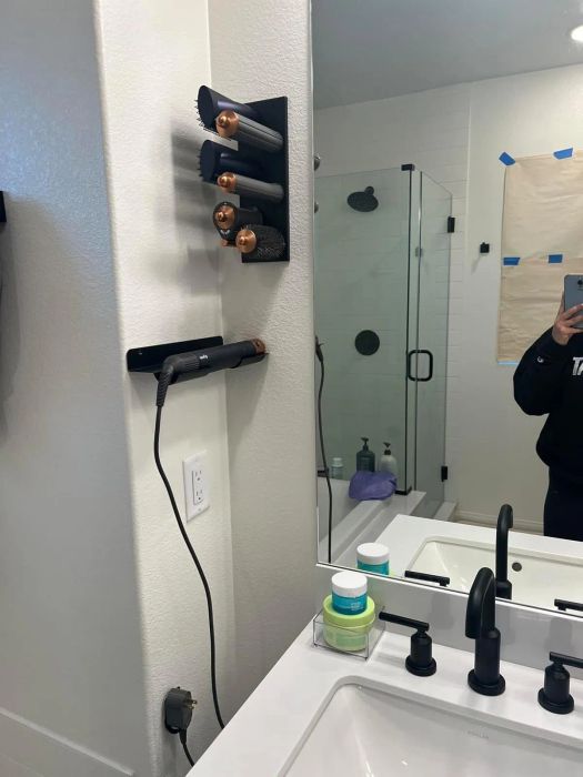 A bathroom counter setup displays a Dyson Airwrap and its accessories, a hair dryer, and personal care products next to a sink. A person's arm can be seen holding up a phone to take the photo