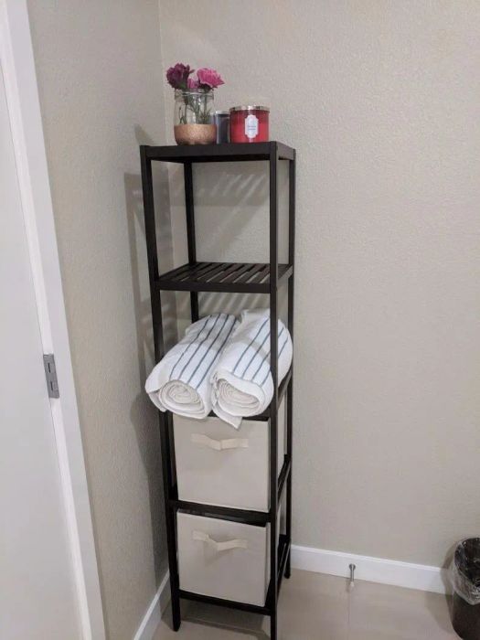 A black metal shelf unit adorned with potted plants, flowers, candles, folded towels, and storage bins, all set against a beige bathroom wall.
