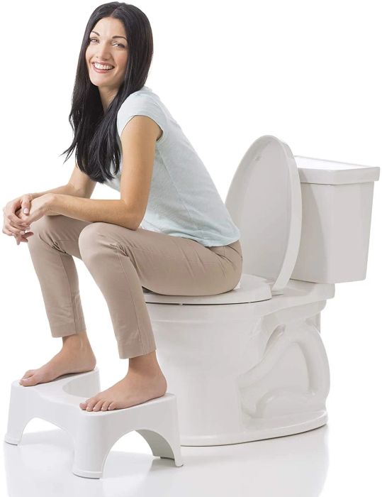 A model sitting on the toilet with their feet comfortably elevated on the white Squatty Potty stool beneath them.