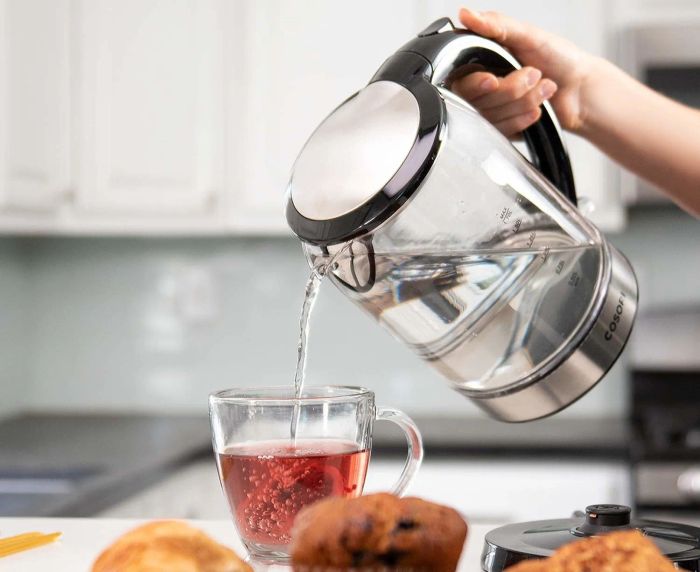A person pouring water from a clear glass electric kettle into a tea cup.