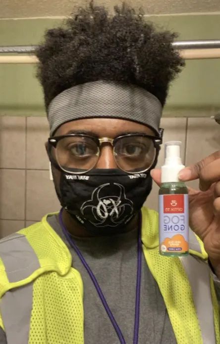 A reviewer photo showing a person wearing clear glasses while holding the anti-fog spray next to their face mask.