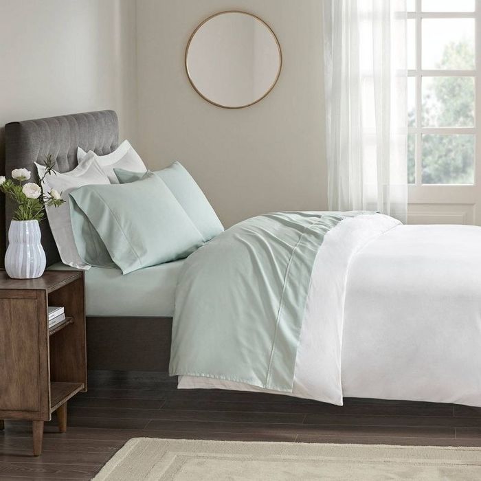 a neatly made bed with green sheets, a nightstand, a rug, and a round mirror hanging on the wall
