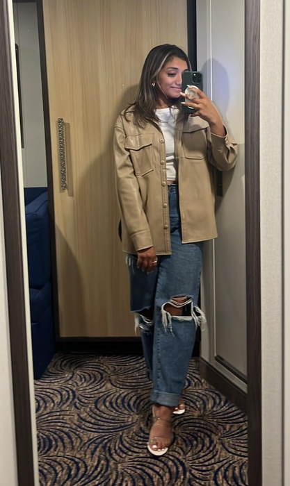 A woman takes a mirror selfie in a casual look: beige jacket, white top, ripped jeans, and white sandals, holding a navy clutch. A perfect outfit for a shopping trip.