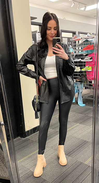 A woman takes a mirror selfie in a store, dressed in a white top, black jacket, black leggings, and beige boots, holding a black handbag.