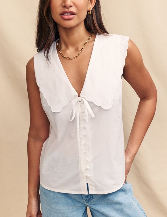 A model wears a sleeveless white blouse with a scalloped collar, a front tie, and blue jeans, posing against a neutral background.