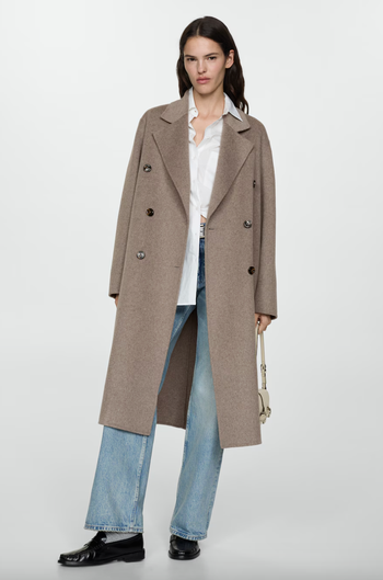 A woman is dressed in a double-breasted trench coat, a white shirt, light-wash jeans, and black shoes, holding a small handbag while standing against a simple background.