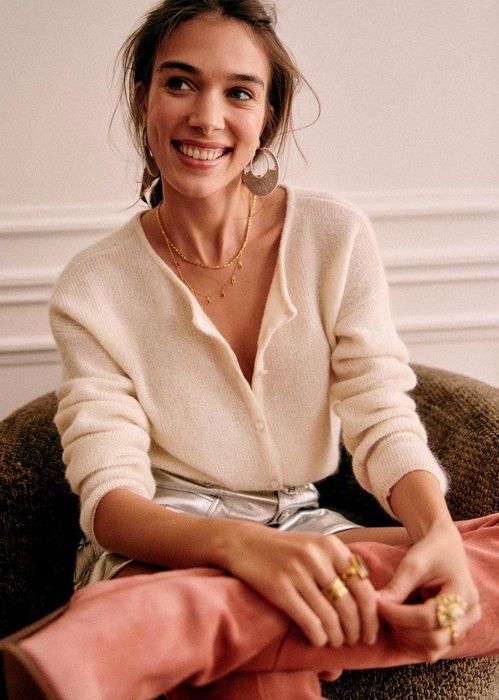 A smiling woman shows off a cozy, button-up sweater paired with metallic pants and hoop earrings, demonstrating stylish everyday fashion for a shopping guide.