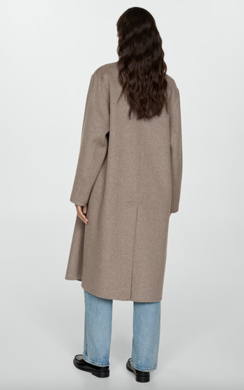 A woman in a long wool coat, blue jeans, and black shoes stands with her back to the camera.