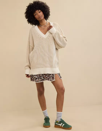 Model rocking an oversized V-neck sweater paired with a leopard print skirt, striped socks, and green sneakers, standing against a simple background