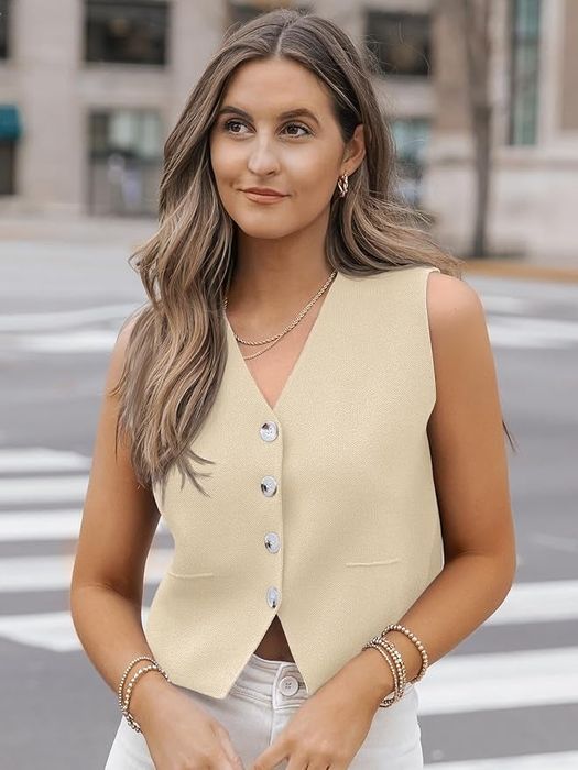 A model stands outdoors, wearing a chic sleeveless beige vest and white pants, accessorized with gold bracelets and necklaces.