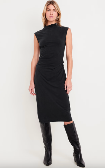 A model flaunts a sleek, sleeveless, knee-length black dress, paired with tall black boots.