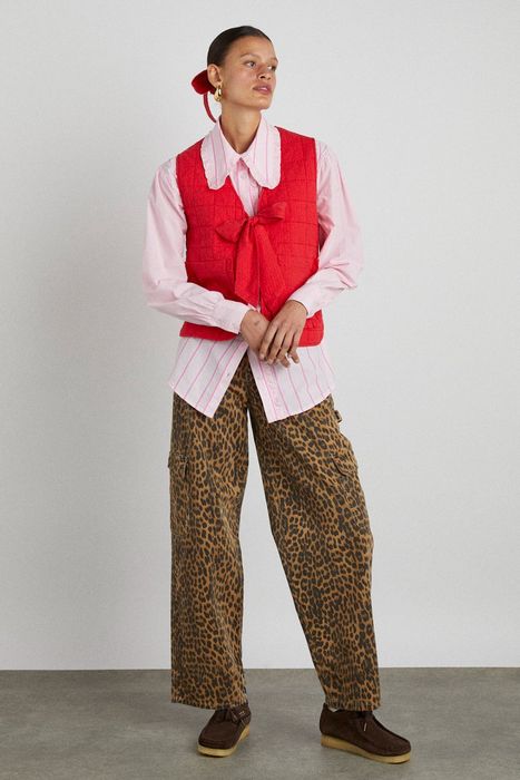 Model wearing a red quilted vest over a pink striped shirt with a large bow tie, paired with leopard print cargo pants and brown shoes for a stylish photoshoot