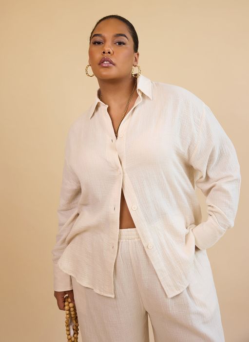 A model wearing a loose-fitting button-up shirt and matching pants, accessorized with large hoop earrings and holding a wooden bead necklace.