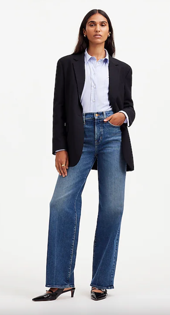 A woman exudes casual chic in a black blazer, light blue shirt, blue jeans, and pointed-toe shoes, standing with one hand tucked into her pocket.