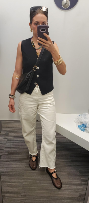 A person in a changing room takes a selfie while wearing a black vest, white pants, dark sandals, and sunglasses, with shopping bags in the background.
