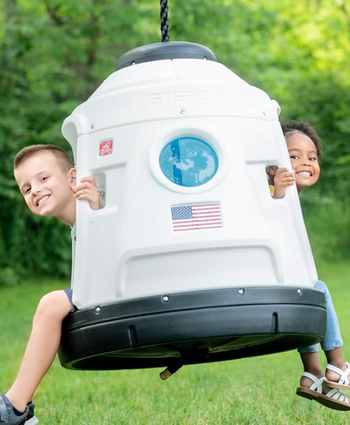 Two children beam with joy while playing on a swing designed to look like a space capsule.