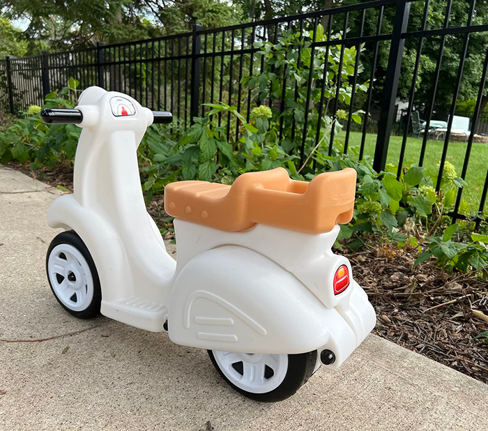 A white and orange toy scooter with three wheels rests on the sidewalk, beside a garden filled with lush greenery.