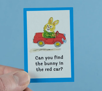 A hand holds a card depicting a bunny riding in a red car with accompanying text.