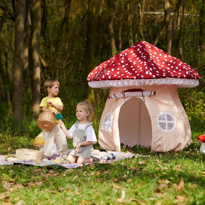 Two children enjoy playing in a forest setting with a mushroom-themed play tent; one holds a basket while the other relaxes on a picnic blanket