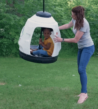 An adult carefully pushes a child seated in a round outdoor swing that mimics the shape of a small rocket.