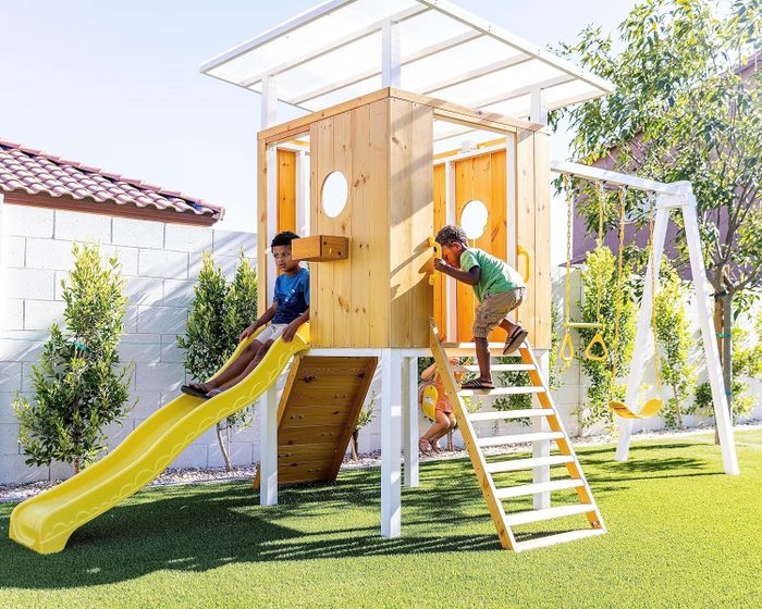 Two children playing on a wooden outdoor playset featuring a slide, climbing wall, swings, and a shaded platform in a backyard.