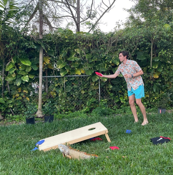 A person prepares to toss a beanbag in a backyard cornhole game, dressed in a patterned shirt and shorts.