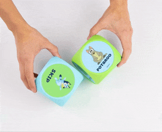 Hands grasp and roll two soft dice, each side displaying animal images and text, featured in an article about shopping items.