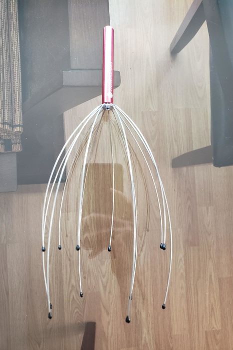 A reviewer’s photo of a metallic pink-handled scalp massager with multiple prongs, each topped with a rubber ball, placed on a table.