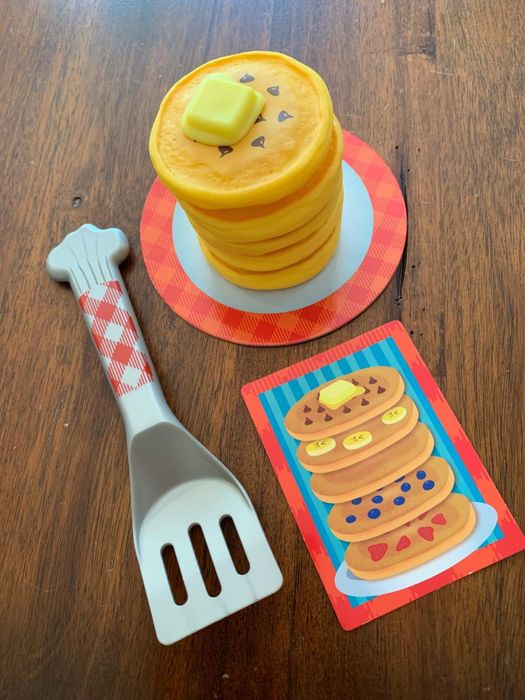 Plastic toy pancakes alongside a spatula placed on a table, with a card beside them featuring three pancakes topped with different ingredients.
