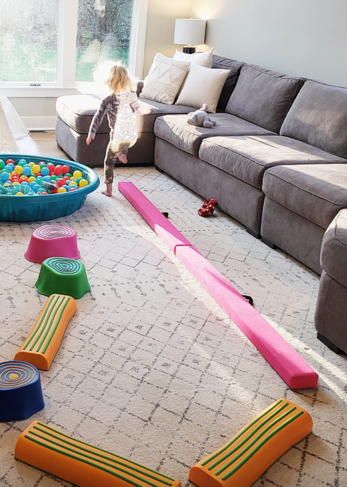 A child navigates a balance beam and stepping stones in a living room, surrounded by a couch, ball pit, and expansive windows.