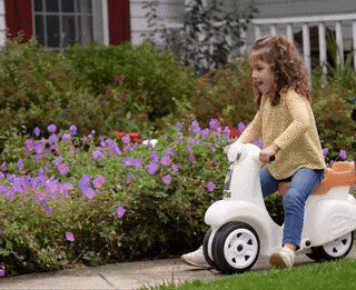 A young child with curly hair rides a white scooter-like toy on the sidewalk in front of a house with a vibrant flower garden.