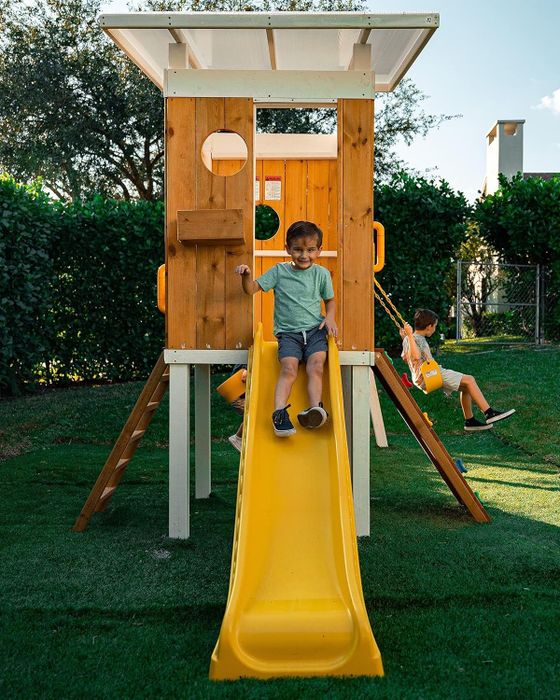 Two children playing on a wooden playset with a yellow slide and swing set in a backyard.