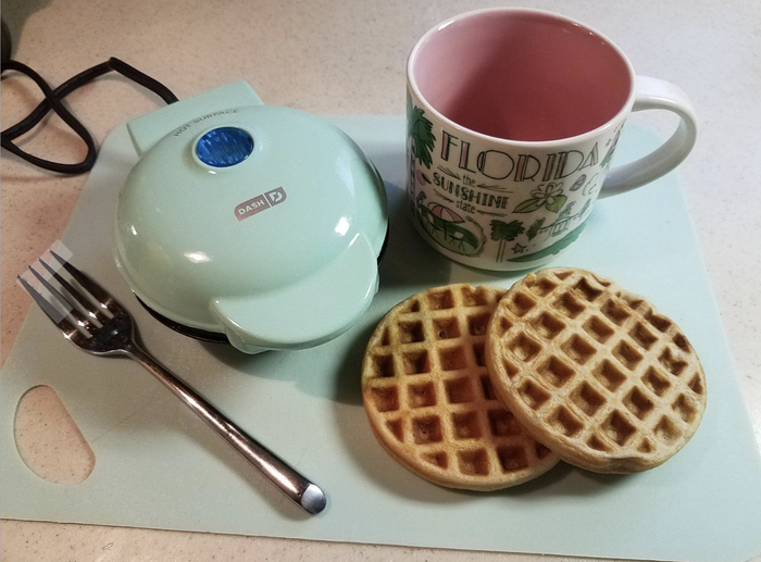 Two golden, cooked waffles next to a small blue waffle maker.
